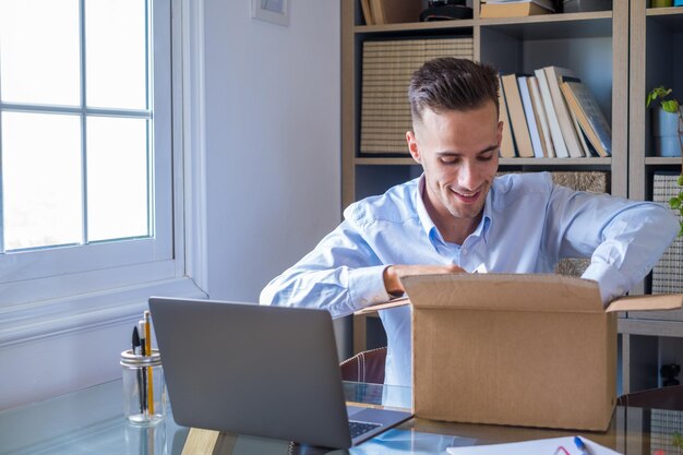 Ein glücklicher lächelnder Mann arbeitet und öffnet eine kleine Schachtel, die gerade zu Hause geliefert wurde, genießt seine Überraschung allein, amüsiert sich, junger kaukasischer Mann, der ein Geschenk oder ein Geschenk im Büro auspackt.