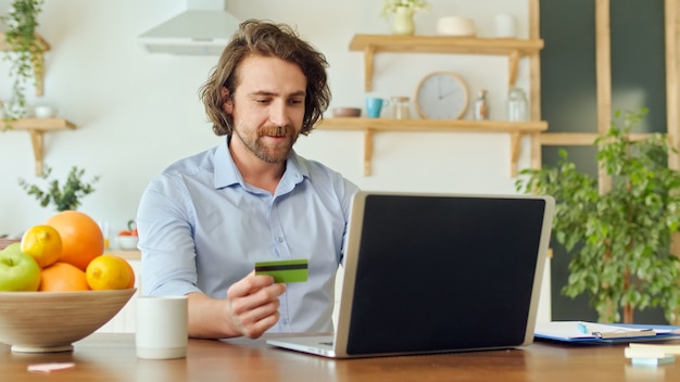 Ein glücklicher Mann benutzt einen Laptop zum Online-Shopping zu Hause und bindet eine Karte für den Online-Shopping-Kauf von t ...