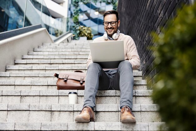 Ein glücklicher Mann sitzt draußen auf der Treppe und benutzt einen Laptop