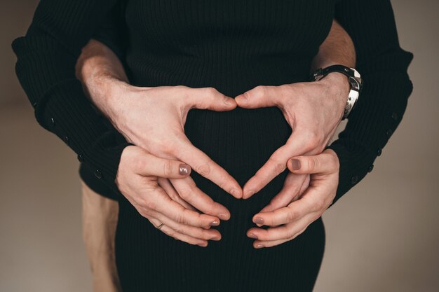 Ein glücklicher Mann steht neben seiner schwangeren Freundin. Ein Mann zeigt einer schwangeren Frau ein Herz aus den Fingern seiner Hände. Ein Mann ist glücklich, Vater zu werden