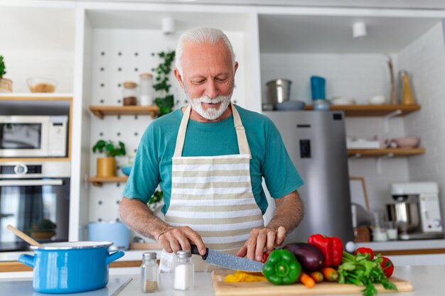 Ein glücklicher pensionierter älterer Mann kocht in der Küche, ein Konzept für pensionierte Hobby-Menschen. Porträt eines lächelnden älteren Mannes, der Gemüse schneidet.