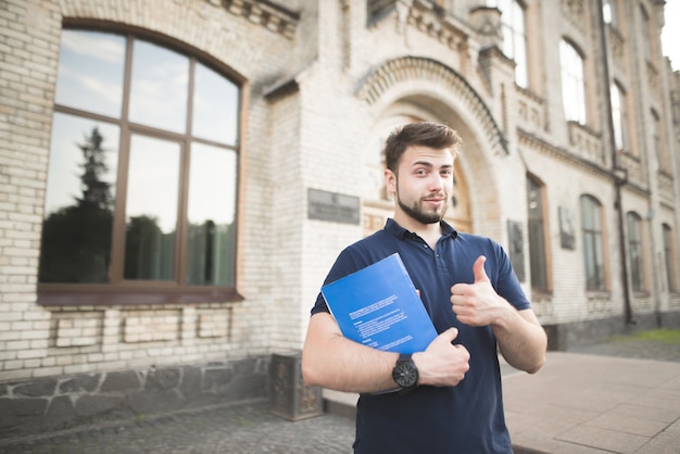 Ein glücklicher Student stolpert vor dem Hintergrund der Universität mit einem Buch und einem Notizbuch in den Händen und zeigt einen Daumen hoch.