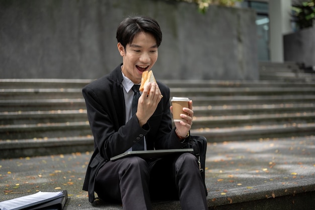 Ein glücklicher und hungriger asiatischer Geschäftsmann isst ein Sandwich auf einer Treppe vor dem Gebäude