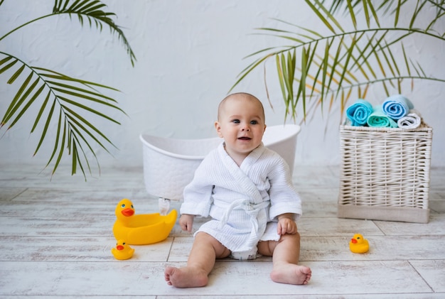 Ein glückliches Baby im Bademantel sitzt mit Entenspielzeug neben einer Babywanne. Kinderhygiene