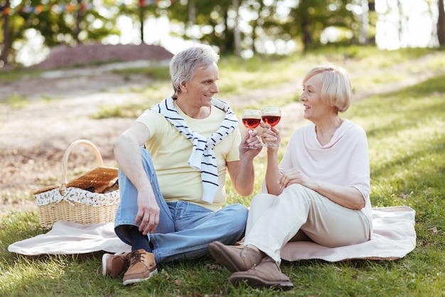 Ein glückliches Ehepaar im Alter, das seine Freude zum Ausdruck bringt, während es ein Picknick genießt und im Park Wein trinkt