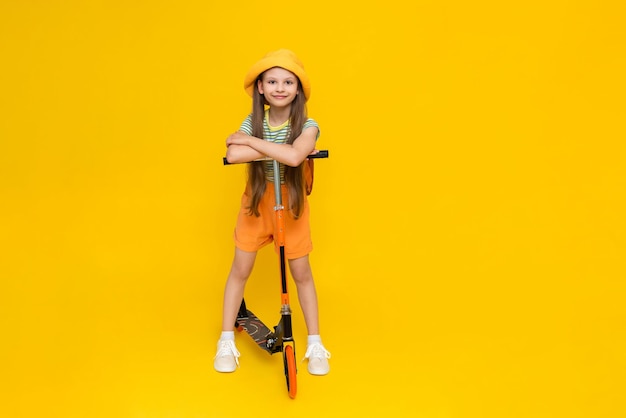 Ein glückliches, fröhliches junges Mädchen in Hut und Shorts, das auf einem zweirädrigen Roller fährt Kinderaktivität in der Stadt Helle gelbe isolierte Hintergrund