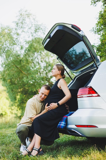 Ein glückliches junges Ehepaar erwartet ein Kind und sitzt im Kofferraum eines Autos in der Natur. Ein Mann und eine Frau umarmen einen schwangeren Bauch, während sie auf einer grünen Wiese sitzen