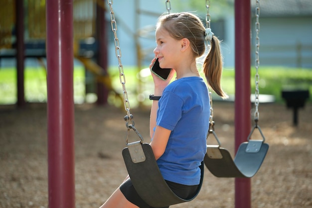 Ein glückliches junges Mädchen spricht im Handy und sitzt im Freien auf Schaukeln im Sommerpark