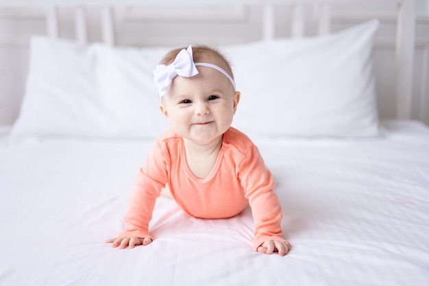 Ein glückliches kaukasisches Mädchen in einem rosafarbenen Bodysuit liegt zu Hause im Schlafzimmer auf weißem Bettzeug auf dem Bett und schaut in die Kamera, lacht ein gesundes Baby