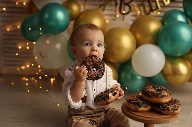 Ein glückliches Kind auf dem Hintergrund von Ballons mit Girlanden isst Schokoladenkrapfen Happy Birthday