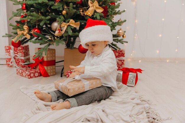 Ein glückliches Kind in einer Weihnachtsmütze sitzt auf dem Boden neben dem Weihnachtsbaum und hält eine Schachtel mit einem Geschenk in den Händen.