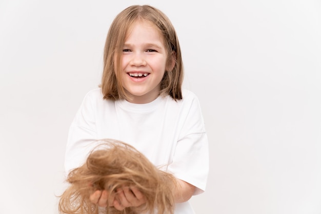 Ein glückliches kindermädchen hält in den händen abgeschnittenes haar, nachdem es auf einem weißen hintergrund geschnitten wurde. bedeutet, kinderhaare zu pflegen. schönheitssalon für kinder.