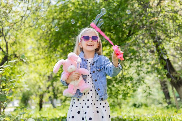 Ein glückliches kleines Mädchen im Sommer mit Sonnenbrille bläst Seifenblasen auf und hält ein Spielzeug in den Händen