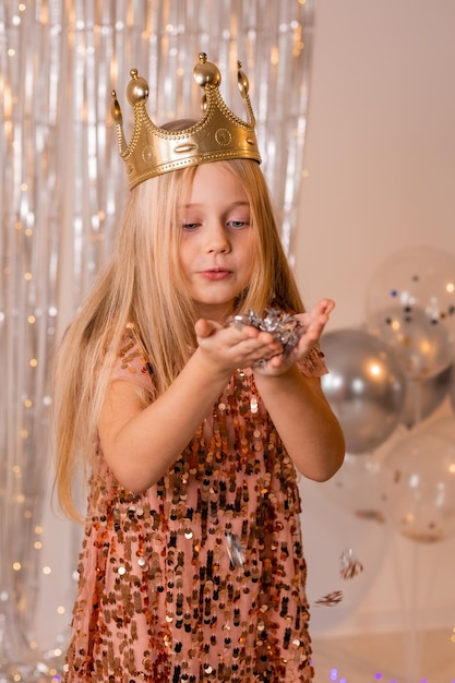 ein glückliches kleines mädchen in einem eleganten kleid und einer krone bläst konfetti von ihren handflächen und wünscht sich etwas.