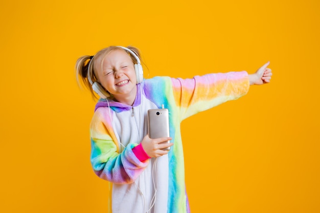 Ein glückliches kleines Mädchen in einem Kigurumi-Einhorn hört Musik in Kopfhörern, die ein Smartphone halten