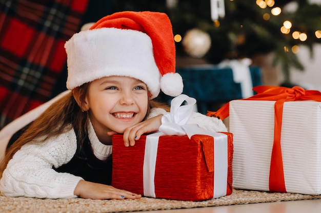 Ein glückliches kleines Mädchen in einem Weihnachtsmann-Hut lächelt mit Geschenken in ihren Händen.