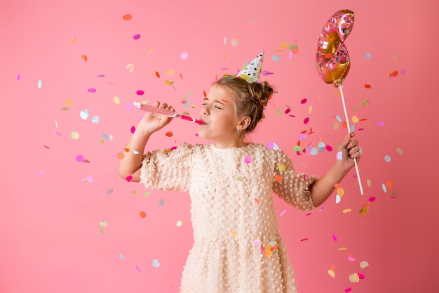 Ein glückliches kleines Mädchen in einer Geburtstagskappe hält einen Donut-förmigen Ballon auf einem rosa Hintergrund im Studio