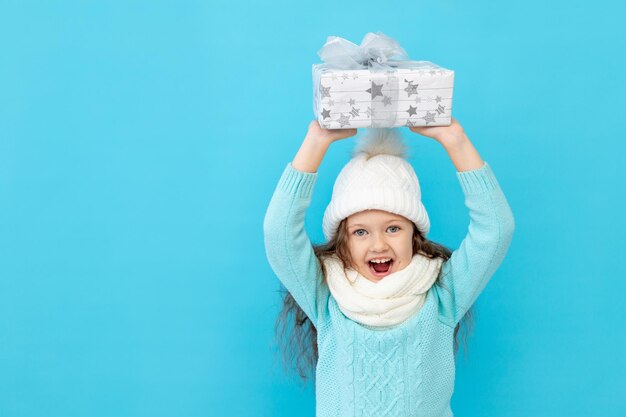 Ein glückliches kleines Mädchen in Winterkleidung, einen Hut und einen Pullover auf blauem, isoliertem Hintergrund hält Geschenke für das neue Jahr oder Weihnachten und lächelt, einen Platz oder Platz für Text