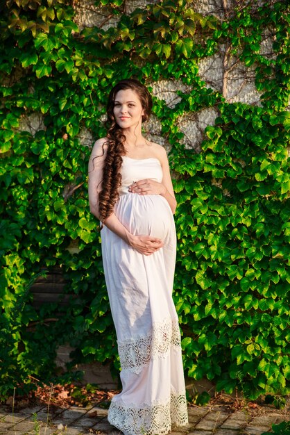 Ein glückliches schwangeres Mädchen in einem weißen Kleid geht im Park spazieren.