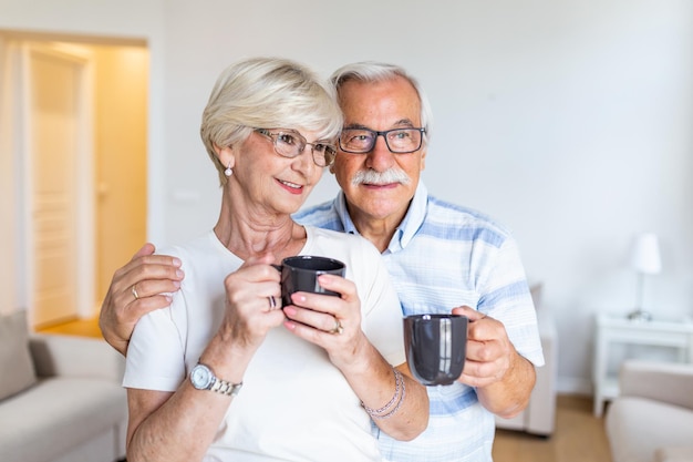 Ein glückliches Seniorenpaar steht im Wohnzimmer, schaut sich gegenseitig an, lächelt und hält eine Tasse Kaffee oder Tee in der Hand, ein älterer Ehemann umarmt seine liebe Frau.