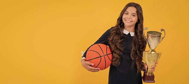 Ein glückliches Teenager-Mädchen hält einen Basketball und einen Champion-Cup auf gelbem Hintergrund.