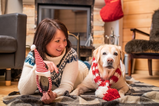 Ein goldener Labrador Retriever in einem Weihnachtsschal liegt mit einer asiatischen Frau mittleren Alters auf einer Decke vor einem Kamin in einem Landhaus.