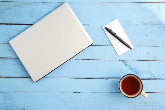 Ein grauer Laptop, eine Tasse Getränk, ein kleines Notizbuch aus Papier und ein schwarzer Stift auf einem blauen Holz