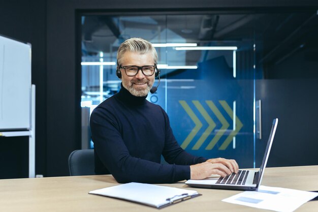 Ein grauhaariger gutaussehender Geschäftsmann mit Kopfhörer und Mikrofon arbeitet an einem Laptop sitzt in der