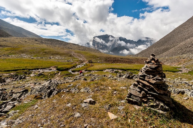 Ein großer Steinhaufen im Vordergrund und große Berge im Hintergrund. Blauer Himmel mit niedrigen weißen Wolken über den Bergen. Mehrere Touristen mit Rucksäcken gehen den Weg entlang. Horizontal.