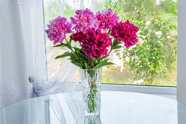 Ein großer Strauß rosa Blumenpfingstrosen steht im Zimmer auf dem Tisch in einer Kristallvase am Fenster