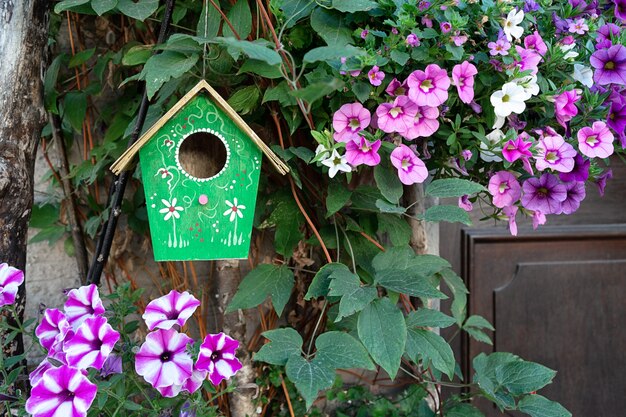 Ein grünes Vogelhaus hängt an einem Baum, der von Petunienblumen umgeben ist.