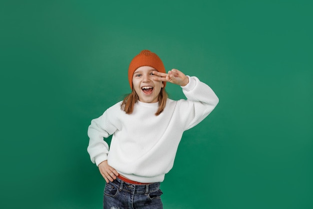 Ein gutaussehender Junge in einem weißen Pullover und Hut lächelt und tanzt und hält seine Finger in der Nähe seiner Augen auf einem isolierten grünen Hintergrund.