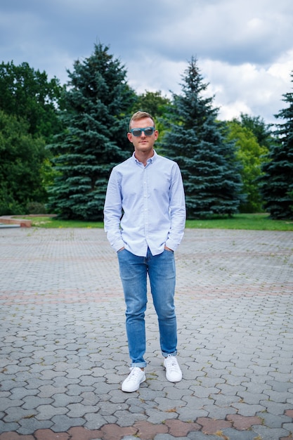 Ein gutaussehender junger Mann europäischen Aussehens mit Sonnenbrille trägt ein Hemd und eine Jeans. Der Typ geht die Straße entlang, er ist stylisch gekleidet