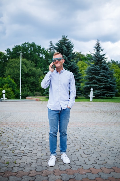 Ein gutaussehender junger Mann europäischen Aussehens mit Sonnenbrille trägt ein Hemd und eine Jeans. Der Typ geht die Straße entlang, er ist stylisch gekleidet