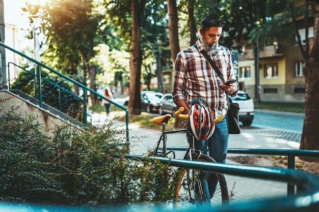 Ein gutaussehender junger Mann fährt mit seinem Fahrrad in die Stadt, geht daneben und liest Nachrichten auf dem Smartphone.