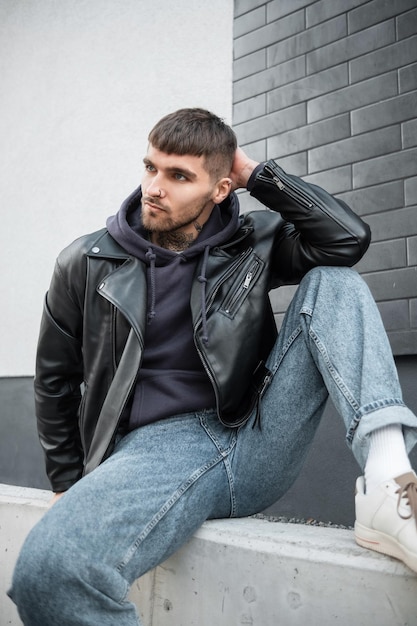 Ein gutaussehender junger Mann, Hipster-Model mit Haaren und einem Bart mit Tätowierung am Hals, der modische Kleidung mit einer Jacke, Hoodie-Jeans und weißen Turnschuhen trägt, sitzt draußen in der Nähe einer schwarzen Ziegelwand