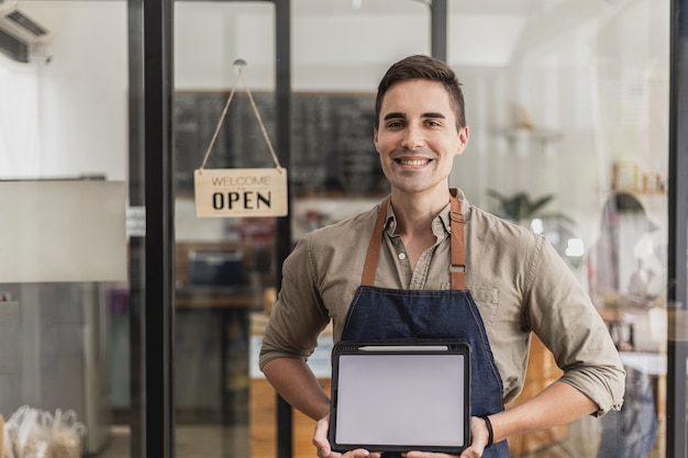 Ein gutaussehender Mann sitzt in einem Café, er hält ein Tablet-Mockup für eine Begrüßungsnachricht oder ein Menü, er ist ein Café-Mitarbeiter mit Schürze und bedient Kunden. Café-Service-Konzept.