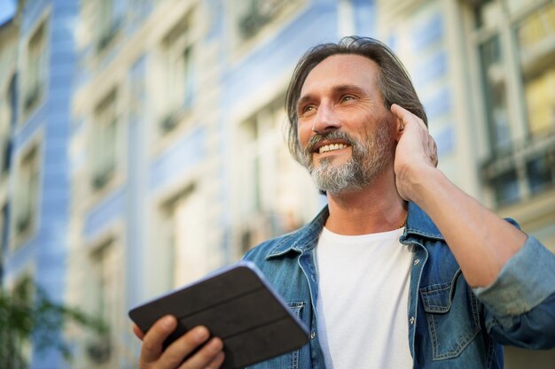 Ein gutaussehender, reisender reifer Mann setzt ein kabelloses Headset ein und nutzt ein digitales Tablet, um im Freien auf den Straßen der Stadt zu telefonieren. Reifer Mann, der Musik hört, reist durch die Straßen der Altstadt