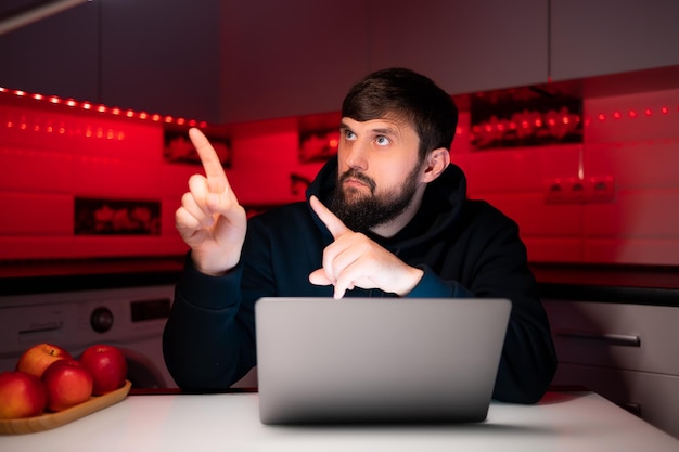 Ein Hacker im schwarzen Hoodie sitzt an einem Laptop und zeigt mit dem Finger zur Seite