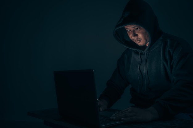 Ein Hacker mit einer Kapuze, die seinen Kopf bedeckt, sitzt mit einem Laptop in einem dunklen Raum