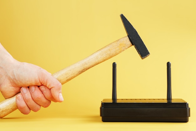 Ein Hammer schlägt auf den WLAN-Modemrouter, um ihn vor einem gelben Hintergrund zu beschädigen.