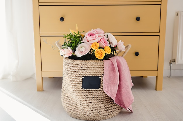 Ein handgefertigter Jutekorb mit einer Kreidetafel zum Beschreiben mit Blumen und einem rosa Handtuch auf dem Boden neben einer gelben Holzkommode mit Schubladen an einer weißen Wand Gemütliches Zuhause