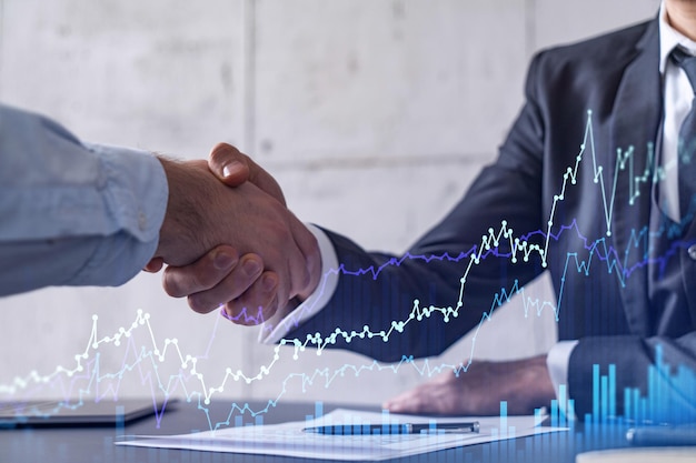 Ein Handshake symbolisiert eine Kapitalmarkttransaktion, um ein profitables Geschäft im Aktienhandel fortzusetzen Finanzhologramm-Diagramm über dem Tisch mit dem Dokument
