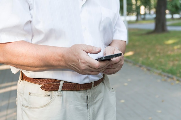 Ein Handy in den großen Händen eines älteren Mannes in weißem Hemd bei einem Spaziergang im Park
