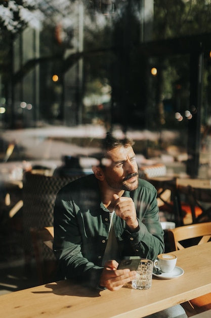 Ein hartnäckiger Mann genießt an einem sonnigen Tag allein Kaffee in einem gemütlichen Café, während er in der Betrachtung am Fenster verloren ist