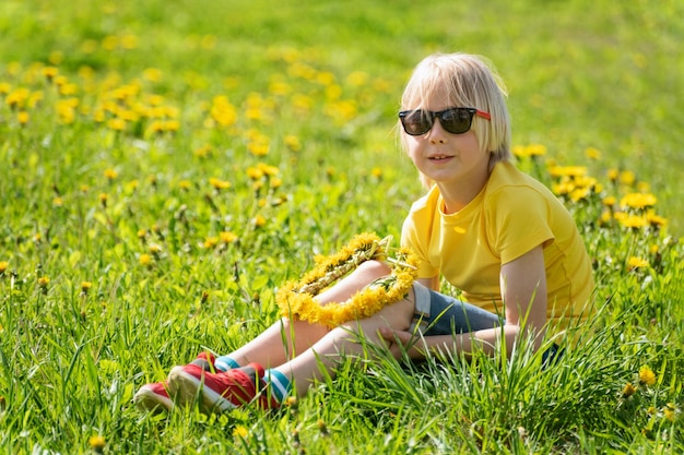 Ein hellhaariges kleines Kind in einem gelben T-Shirt sitzt auf dem grünen Rasen. Ein Kind webt einen Kranz aus Löwenzahn, der auf dem Feld sitzt.