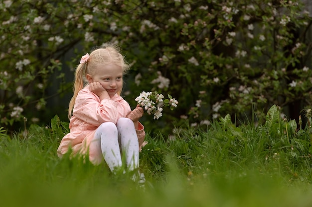 Ein hellhaariges Mädchen von 45 Jahren in rosa Kleidung sitzt auf grünem Gras mit Blumen und schaut nachdenklich in die Ferne. Kind im blühenden Garten