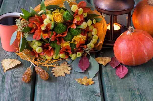 ein herbststrauß aus herbstblumen und rosen in gelb- und orangetönen, herbstlaub, einer laterne mit kerze, einer tasse tee und zwei kürbissen