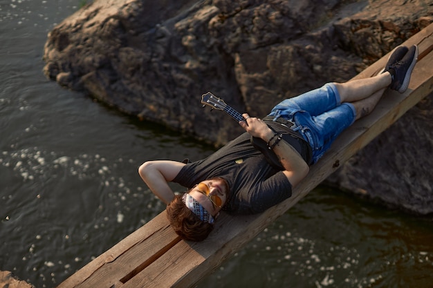 Foto ein hippie-mann mit brille und taschentuch auf dem kopf liegt auf einer holzbrücke über dem fluss und ...