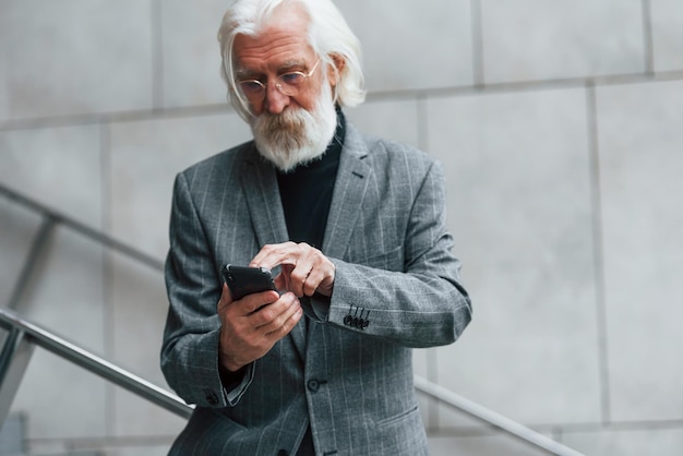 Ein hochrangiger Geschäftsmann in formeller Kleidung mit grauem Haar und Bart versucht im Freien drahtlose Kopfhörer mit Telefon